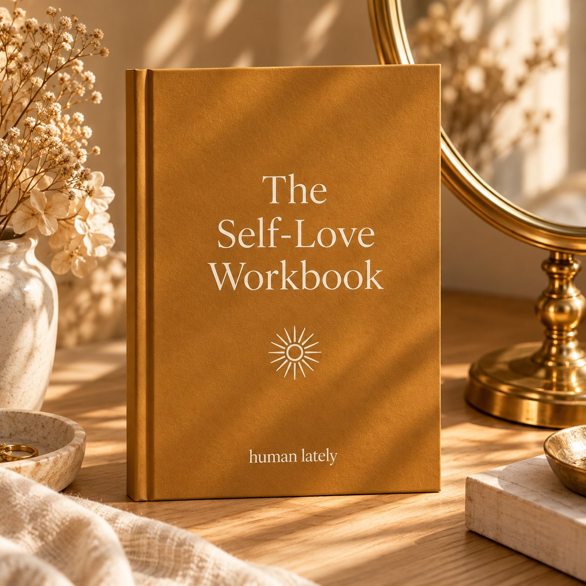 An ochre workbook titled The Self-Love Workbook standing beside a brass mirror